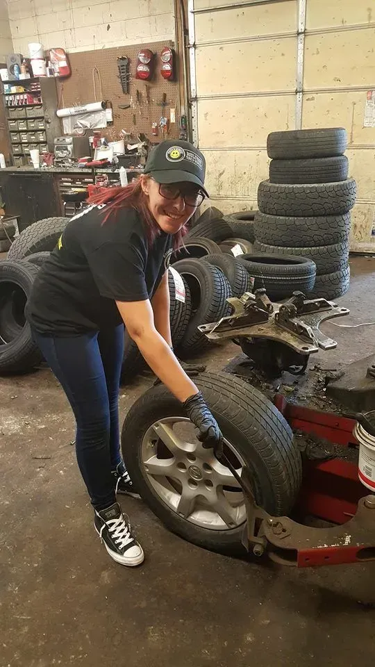 Person in a tire shop lifting a car tire onto a wheel balancing machine with stacked tires in the background