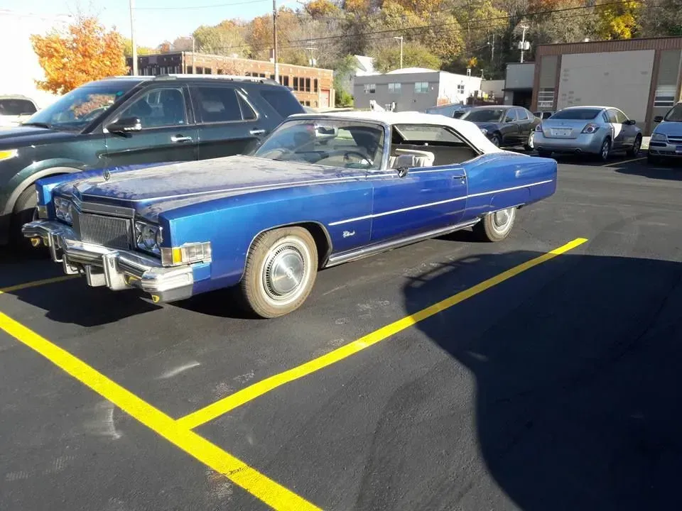 Blue classic convertible car parked in a lot with other vehicles and autumn trees in the background