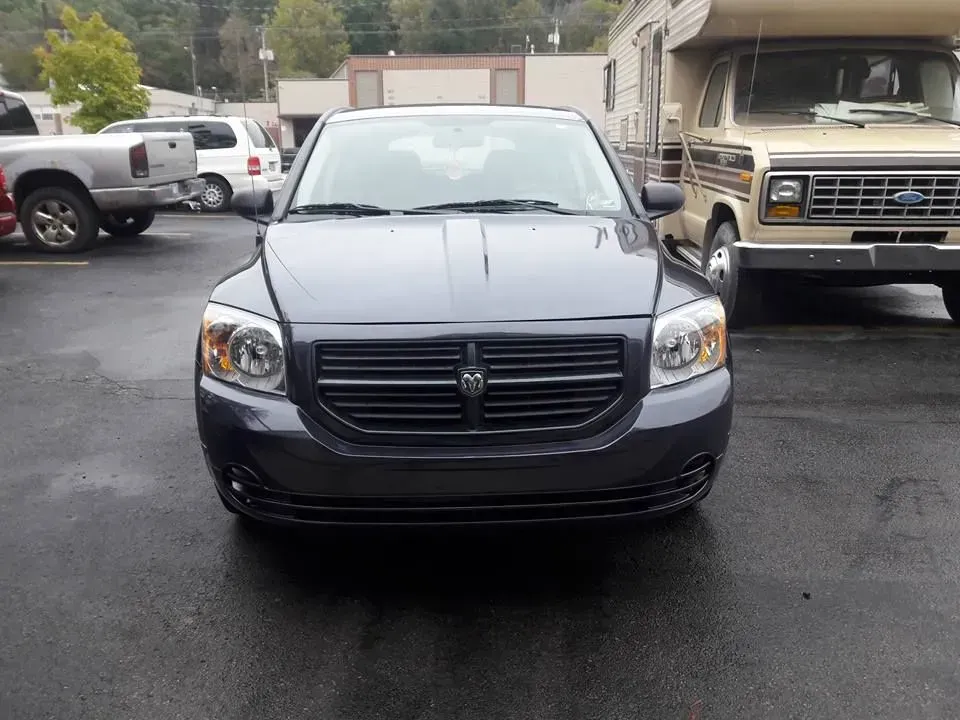 Front view of a dark gray sedan parked in a lot, with trucks and buildings in the background.