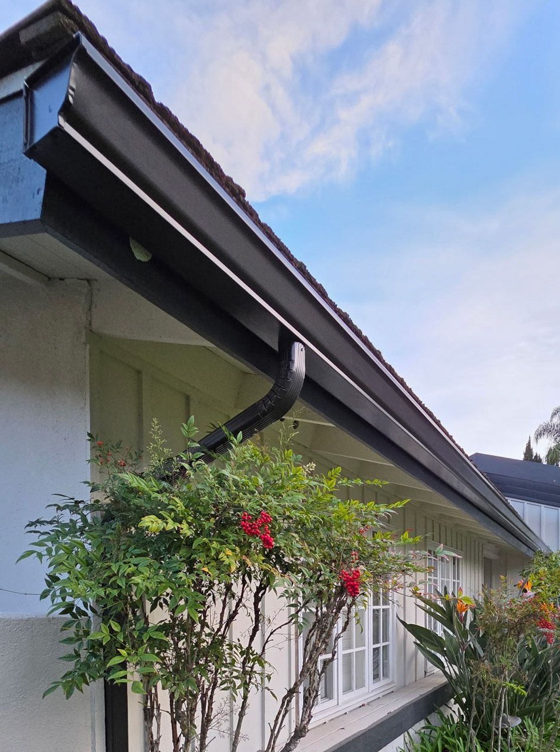 Black gutters on a white building, with bushes and a partly cloudy blue sky. New seamless gutters installed by Gutter Specialist in Reno, NV