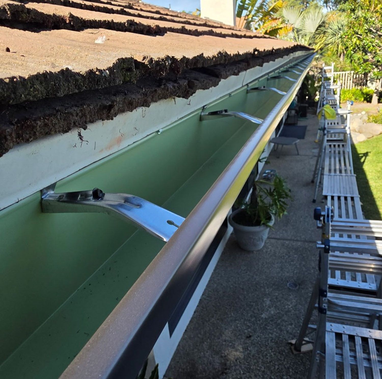 Green rain gutter with metal supports attached to roof edge. Ladder and scaffolding on concrete. New seamless gutters installed by Gutter Specialist in Reno, NV