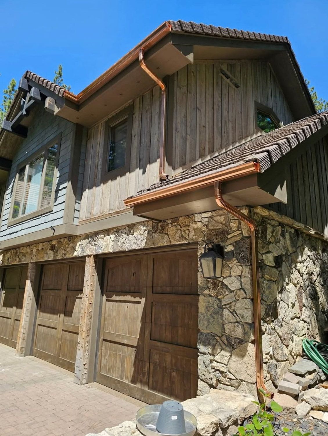 Rustic two-story house with stone base, wooden garage doors, and copper gutters under a sunny sky. New seamless gutters installed by Gutter Specialist in Reno, NV