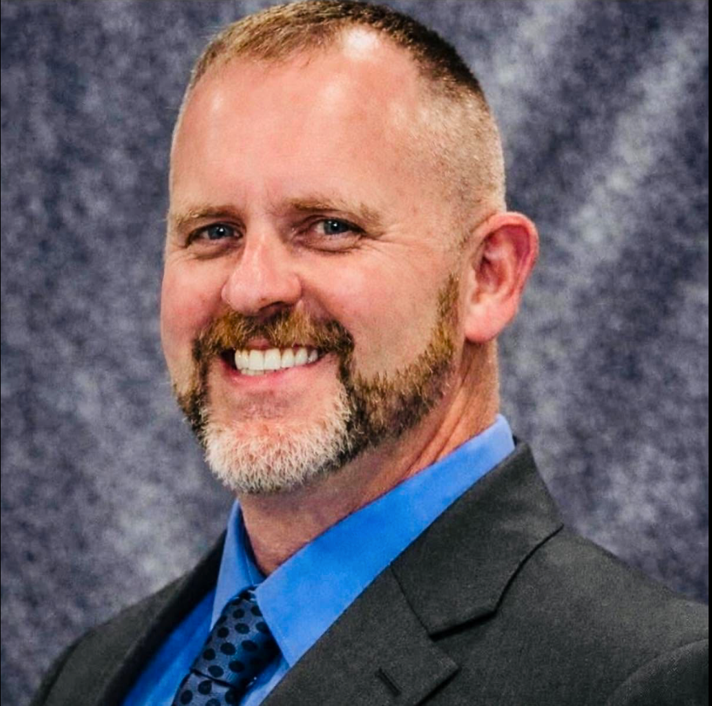 Smiling man in a dark suit and blue shirt against a mottled gray studio backdrop