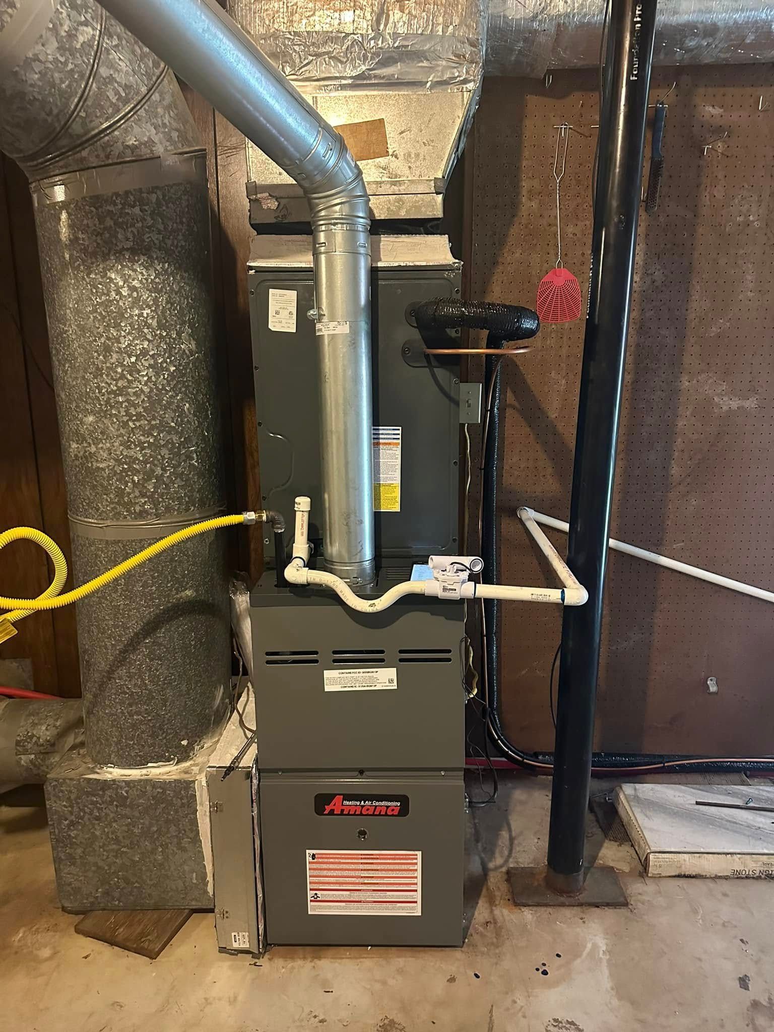 A gray Amana furnace and attached ductwork stand in a basement, with connected metal and white PVC piping.