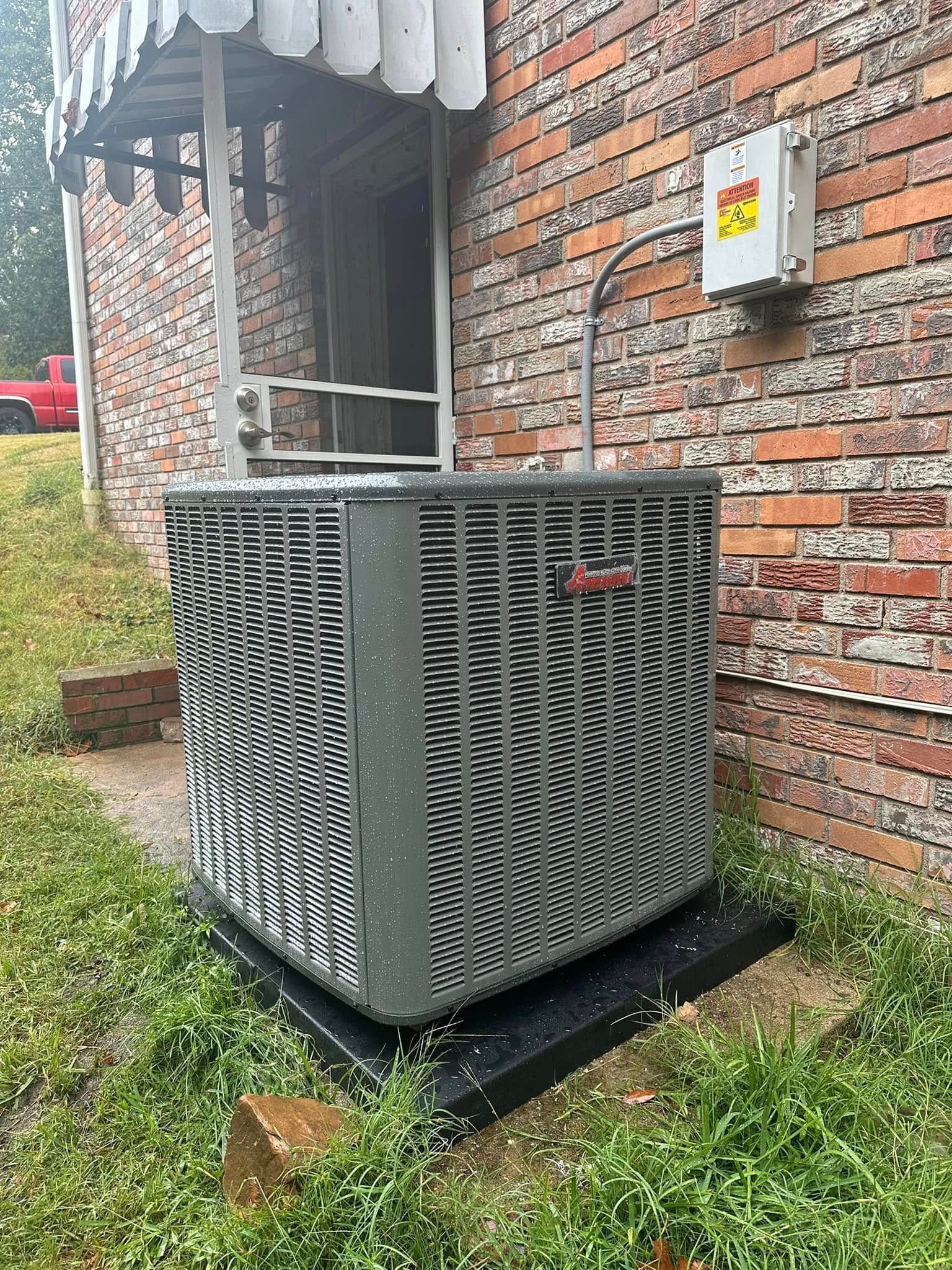 A grey air conditioning condenser unit sits on a black pad against a brick house wall near a porch.