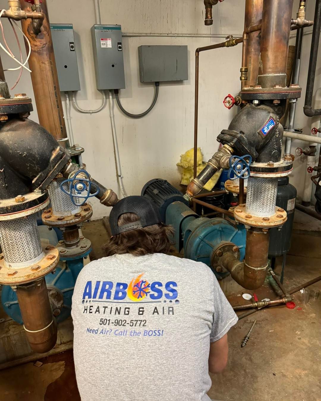 A service technician in an Airboss Heating & Air shirt works on industrial pipes and a pump motor in a utility room.