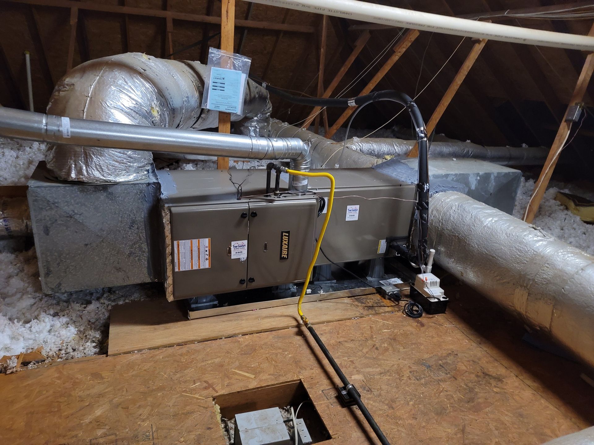 An HVAC furnace unit with ductwork and a yellow gas line installed in an attic with wooden flooring.