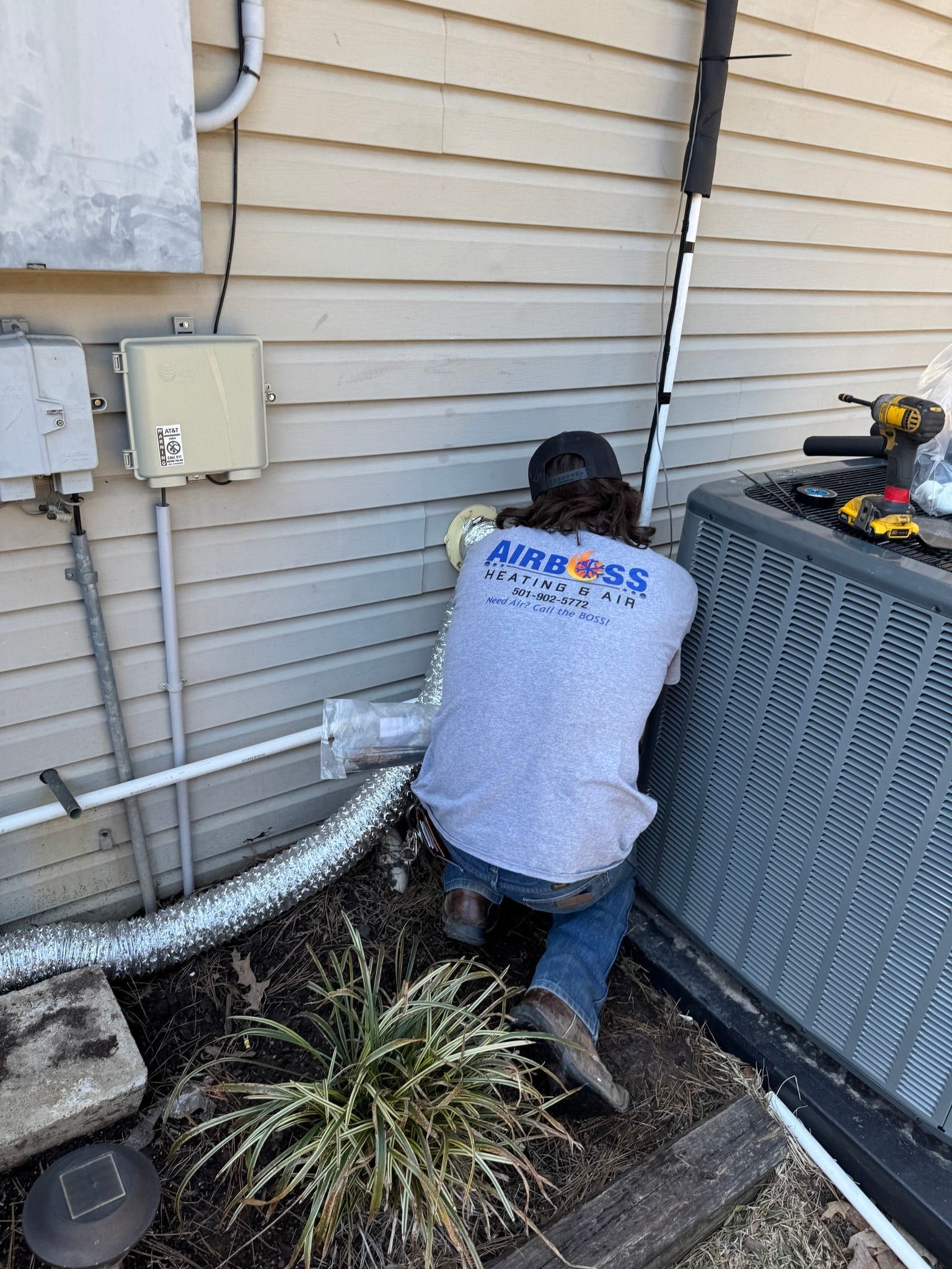 A technician in a gray shirt works on an outdoor HVAC unit against the siding of a house.