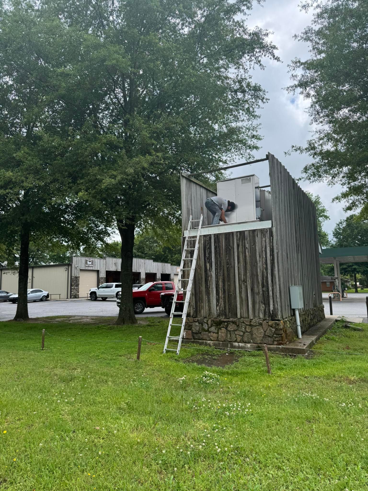A person stands on a ladder against a tall, rectangular wooden structure with a stone base in an outdoor, grassy area.