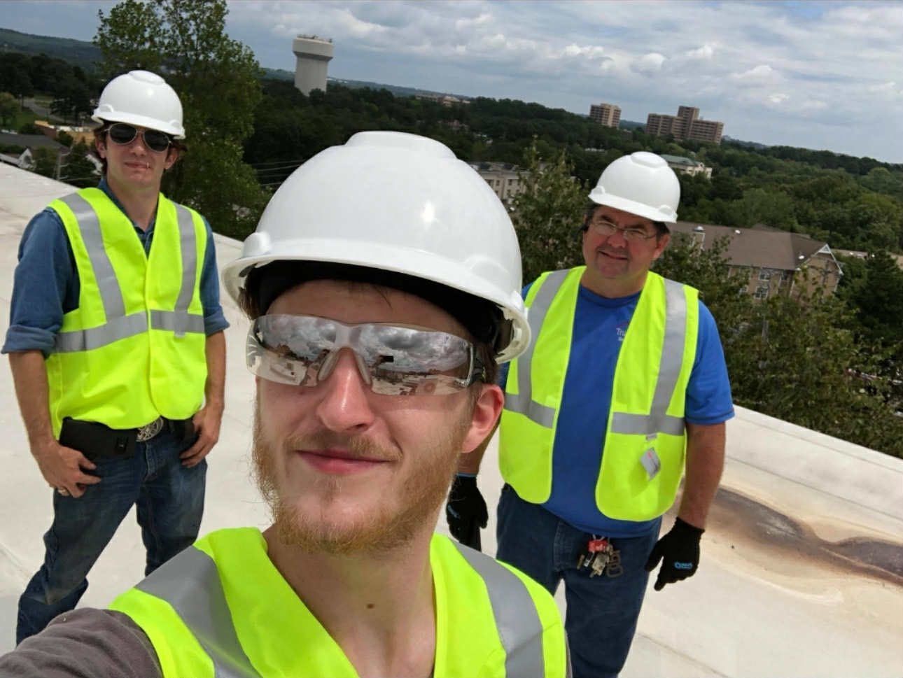 Three people in safety gear, hard hats, and reflective vests taking a selfie on a flat roof overlooking a landscape.