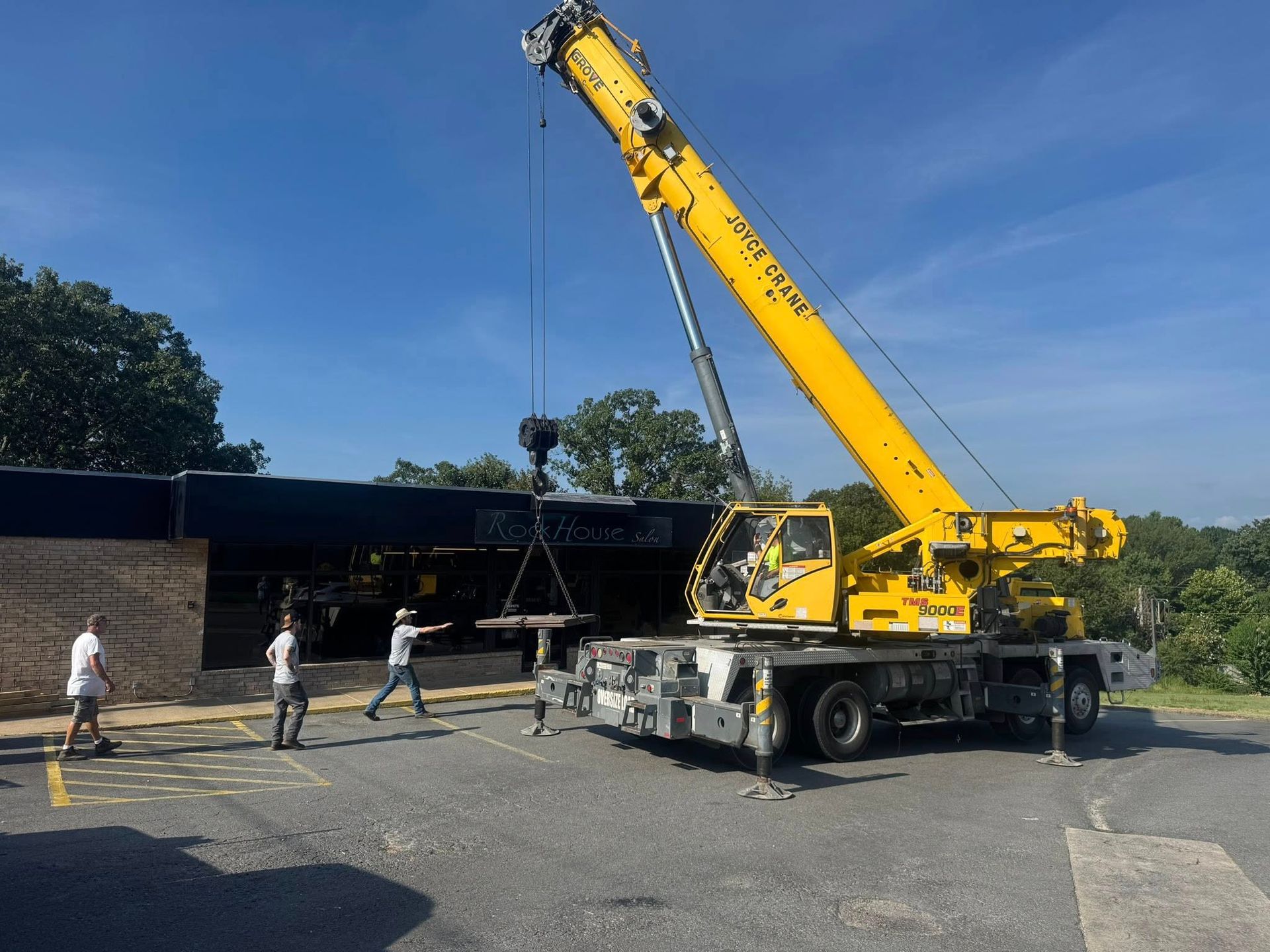 A yellow mobile crane parked on an asphalt lot lifts a circular load, while two people stand nearby on a sunny day.