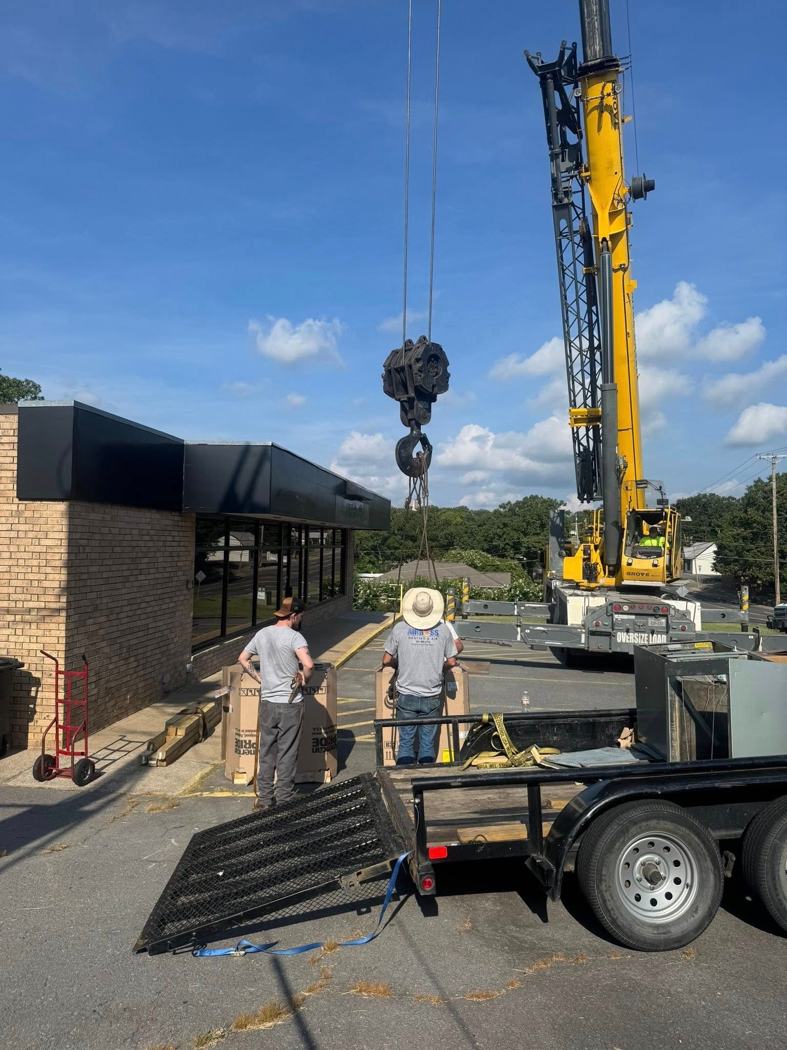 A large yellow construction crane hoists a load near a brick commercial building as two workers stand by a trailer.