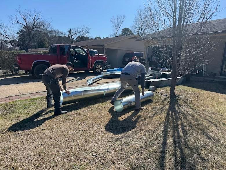 Two workers handle long metal ductwork pipes in a residential yard near a parked red truck.