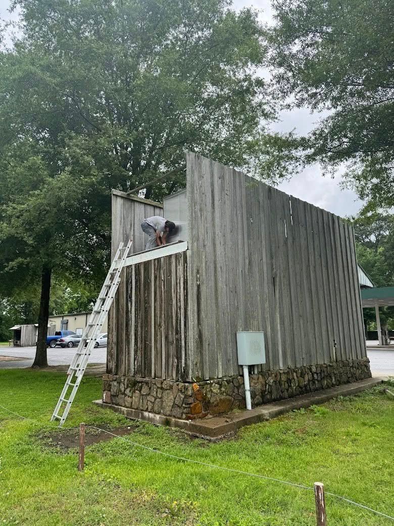 A person climbs a ladder leaned against the tall, weathered wooden wall of a building set in a grassy, tree-filled lot.