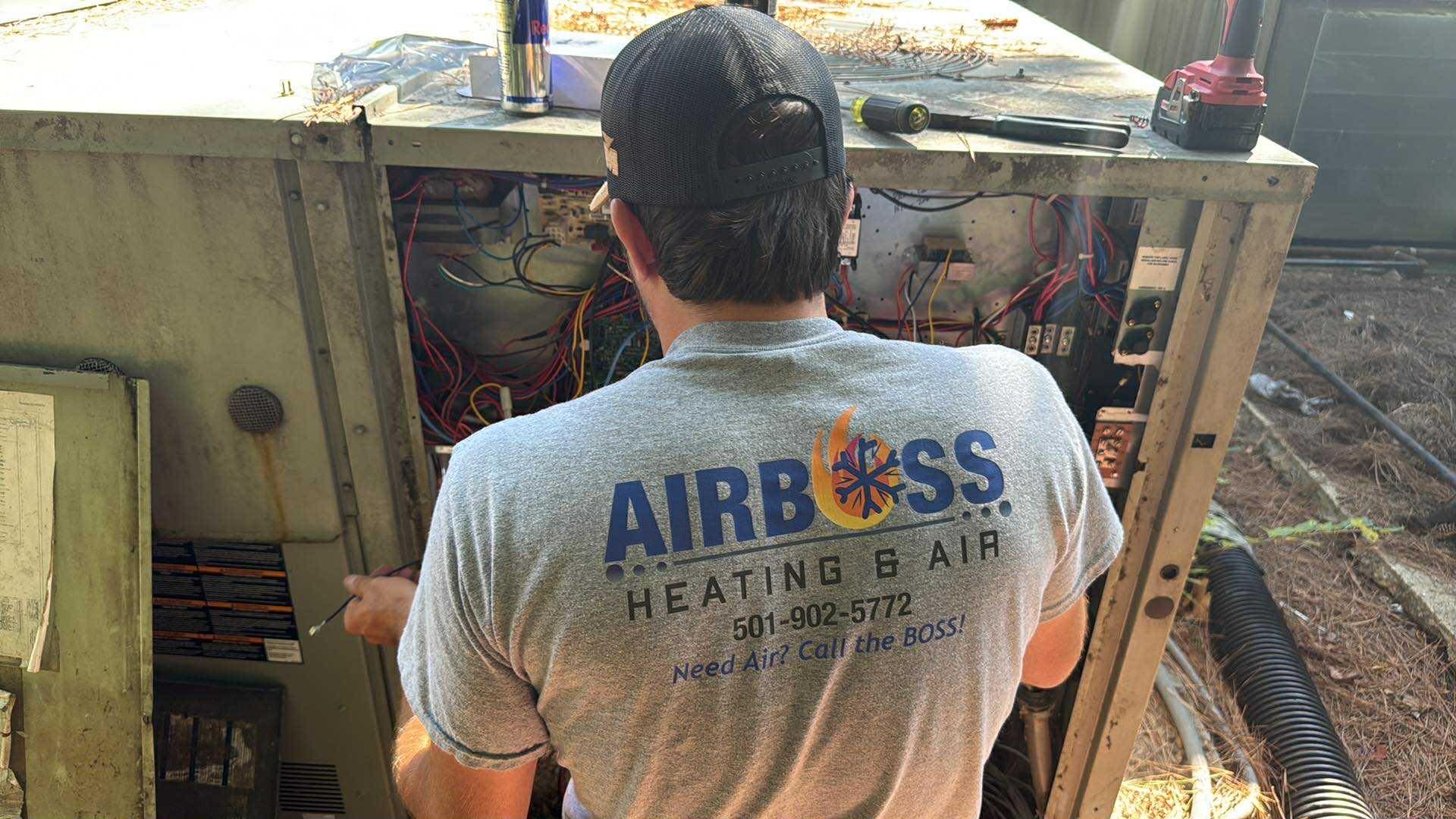 A technician in a gray Airboss Heating & Air t-shirt repairs the electrical components of an outdoor HVAC unit.