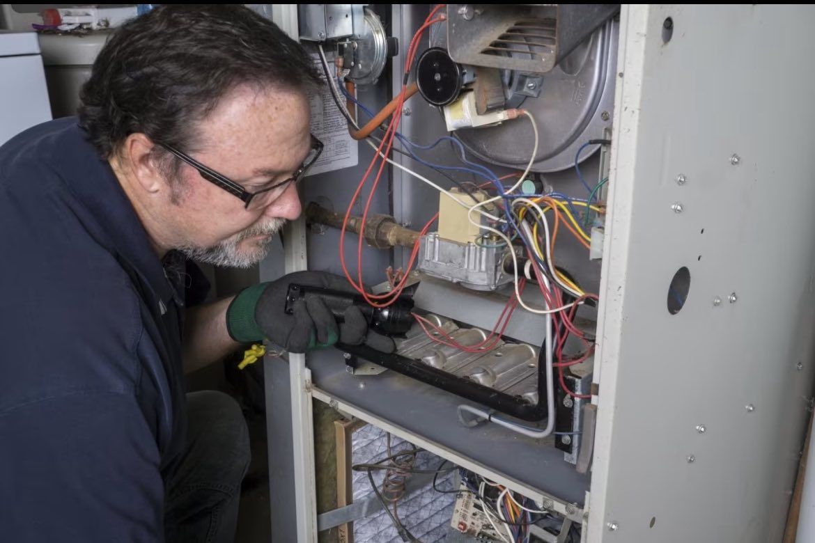A technician wearing glasses and gloves inspects the internal wiring and components of an open furnace unit.