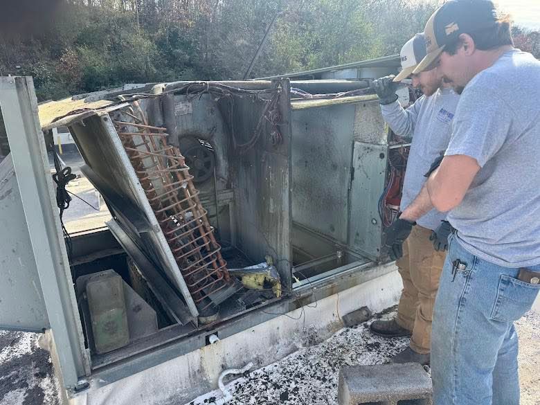 Two people in work clothes inspect the open interior of a rooftop commercial HVAC unit on a sunny day.