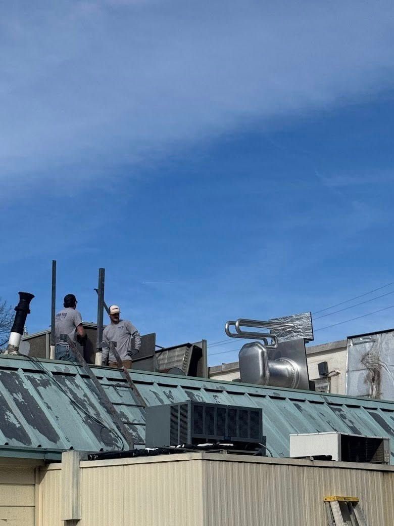 Two workers in hard hats install equipment on a green metal roof under a bright blue sky.