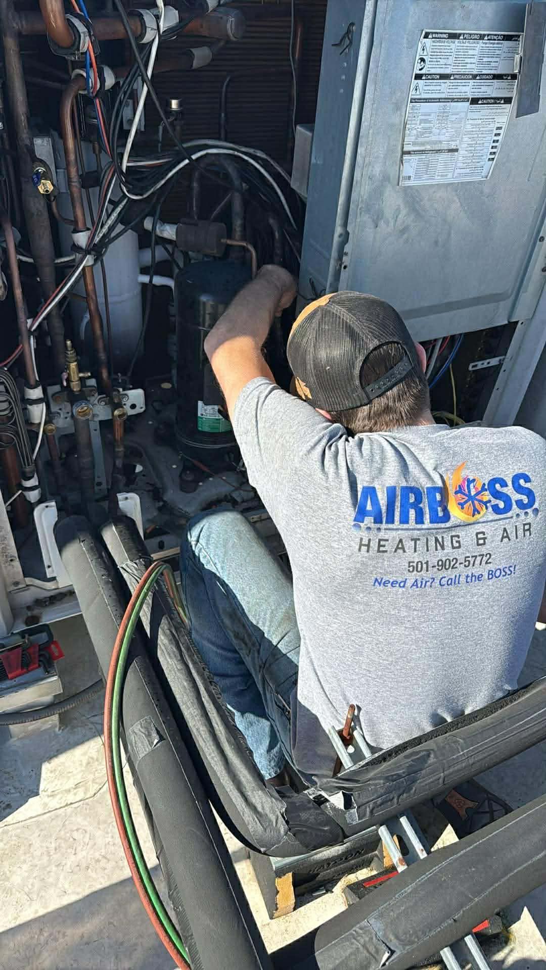 A technician in an Airboss Heating & Air shirt repairs the internal components of an outdoor HVAC unit.
