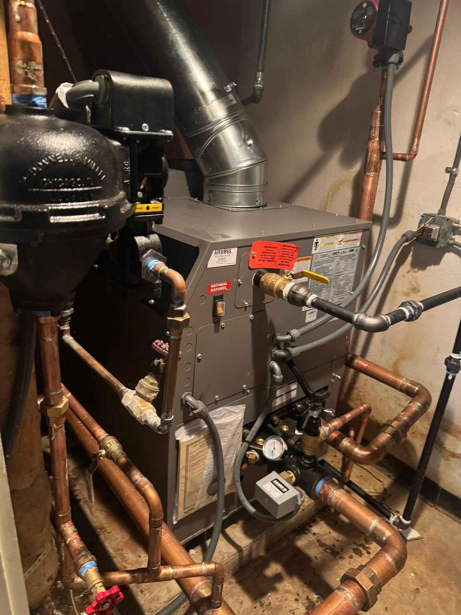 A grey boiler system in a basement with various copper pipes, a flue pipe, and electrical connections.