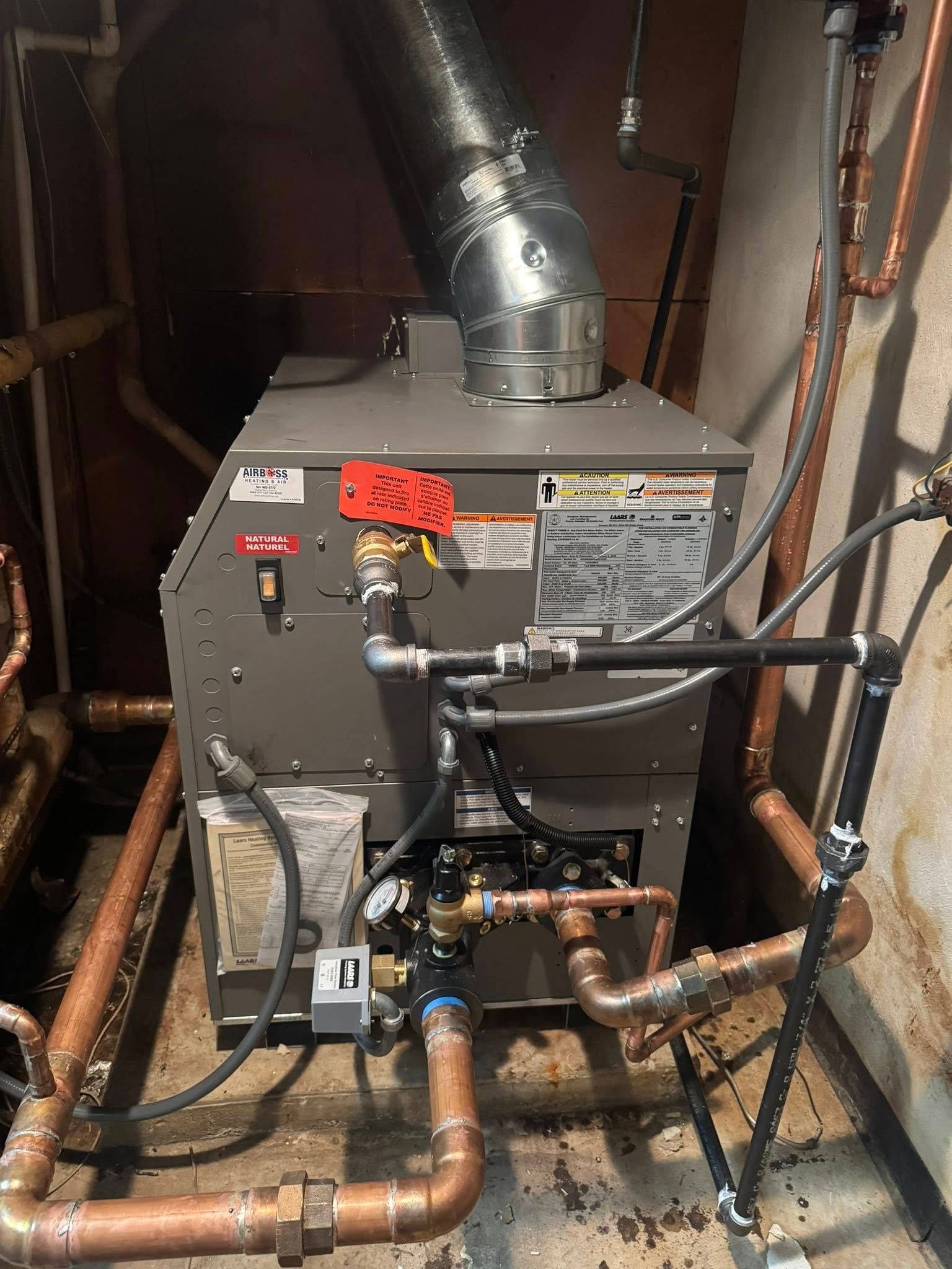 A grey, industrial boiler with copper pipes and a metal exhaust vent in a dimly lit basement.