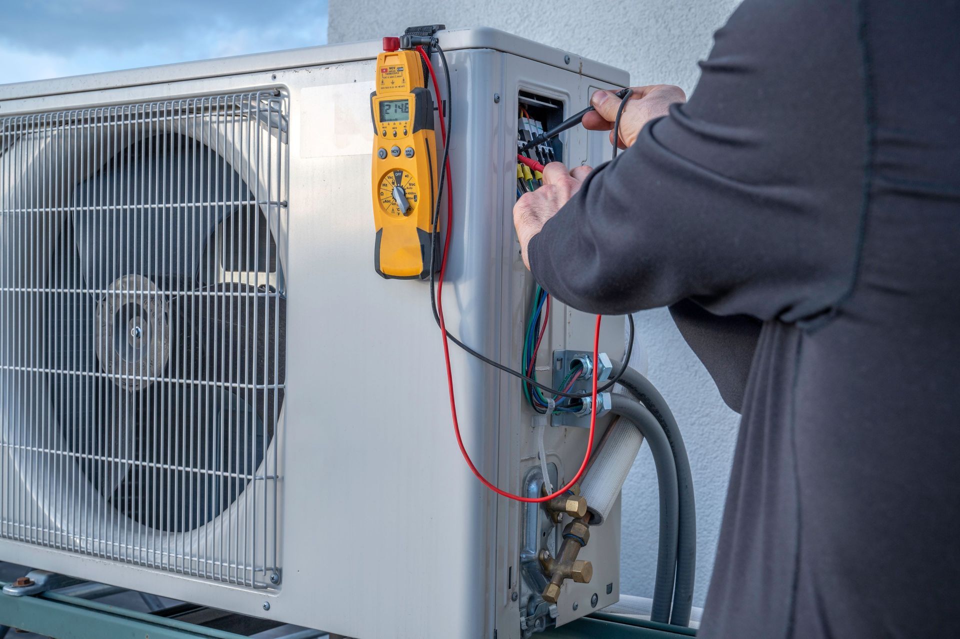 A person in a gray shirt uses a yellow multimeter to test the electrical wiring of an outdoor HVAC unit.