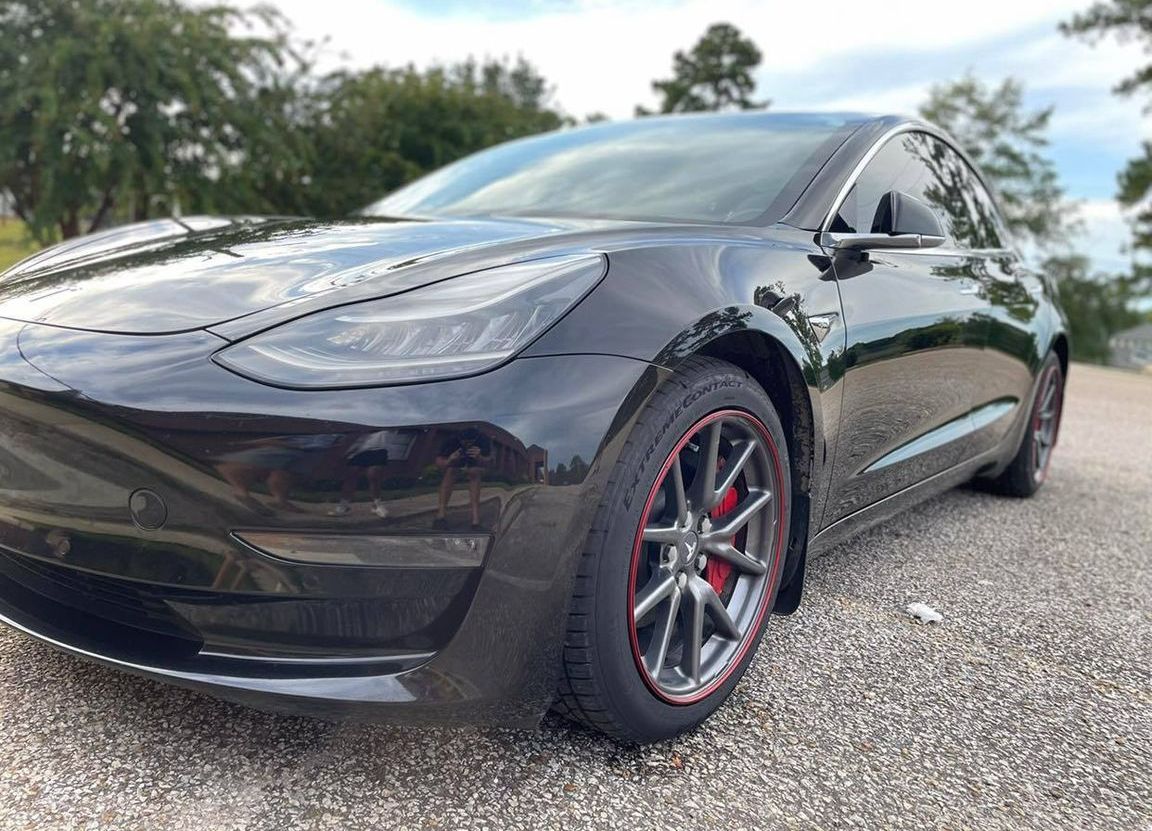 Black Tesla sedan parked on gravel, low-angle front-left view with red-accented wheels.