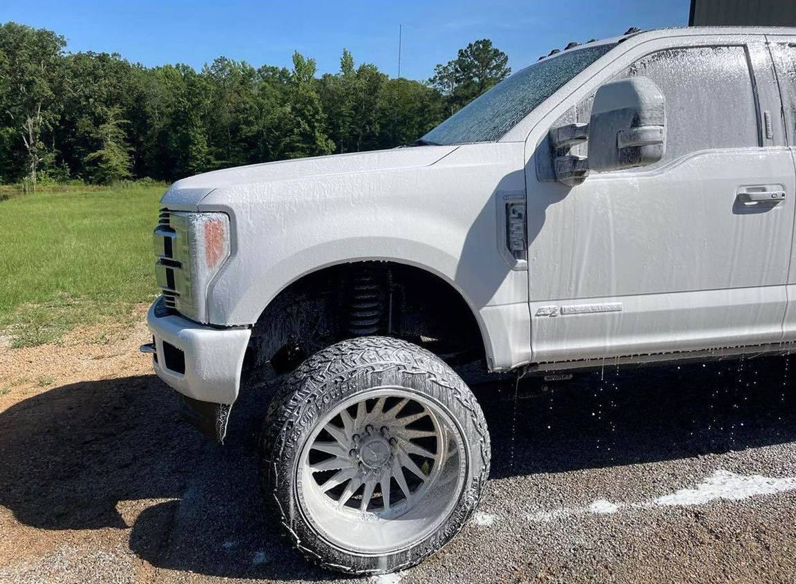White pickup truck being washed outdoors, covered in soap suds, with trees and grass in the background.