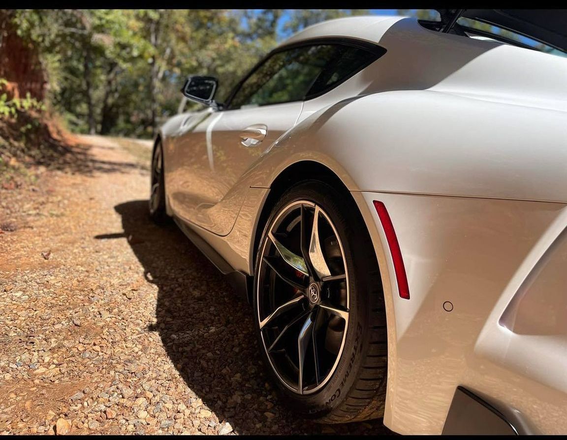 White sports car parked on a dirt road beside trees, viewed from the rear side.