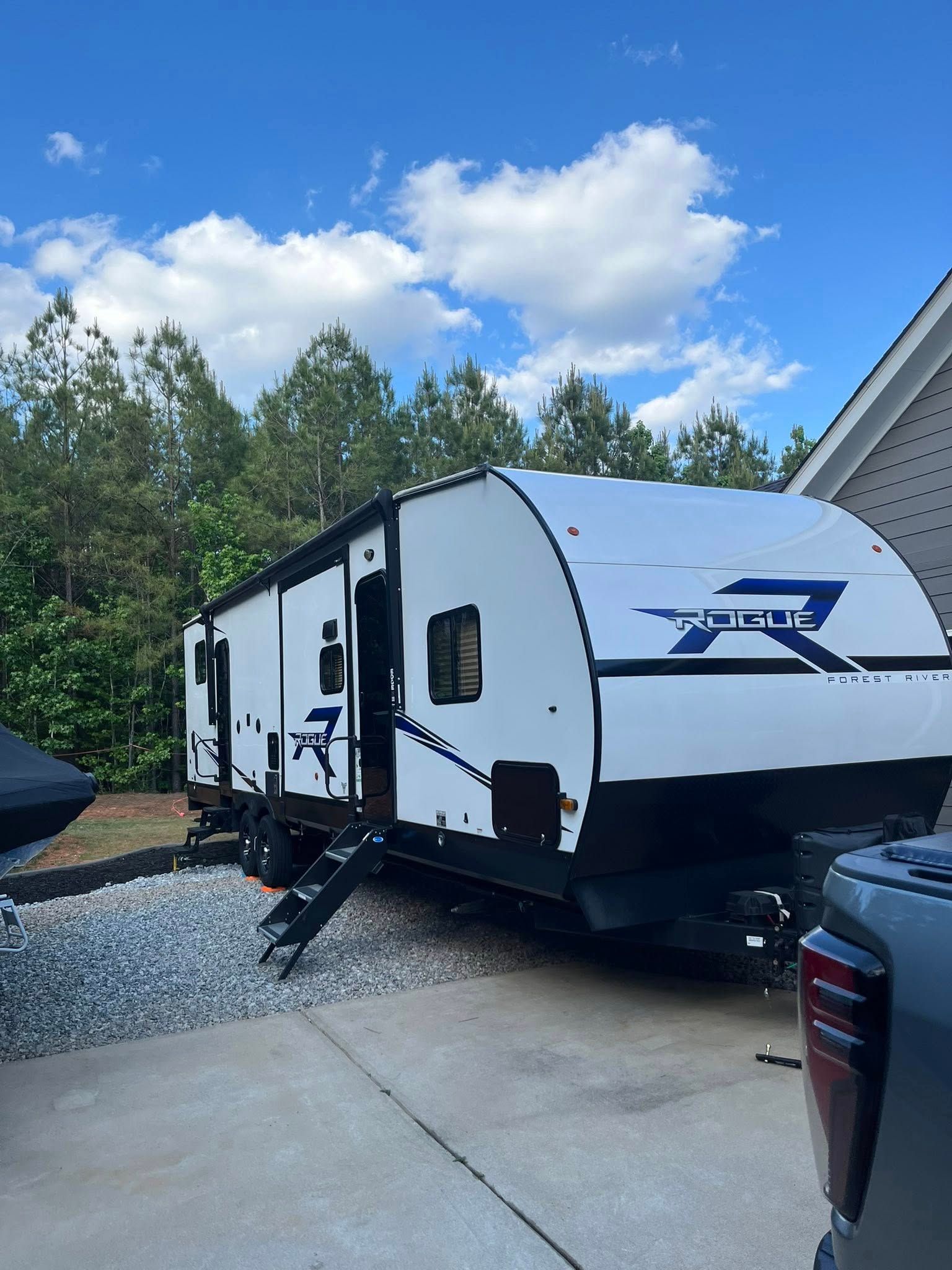 White and black travel trailer parked on a driveway under a blue sky with trees nearby