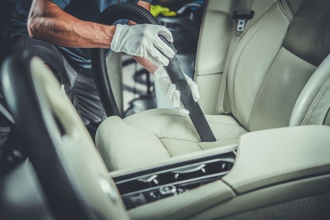 Person vacuuming a beige car seat and floor with a handheld vacuum in a car interior