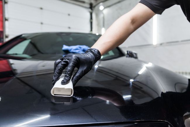 Gloved hand polishing a black car hood with a sponge in a garage