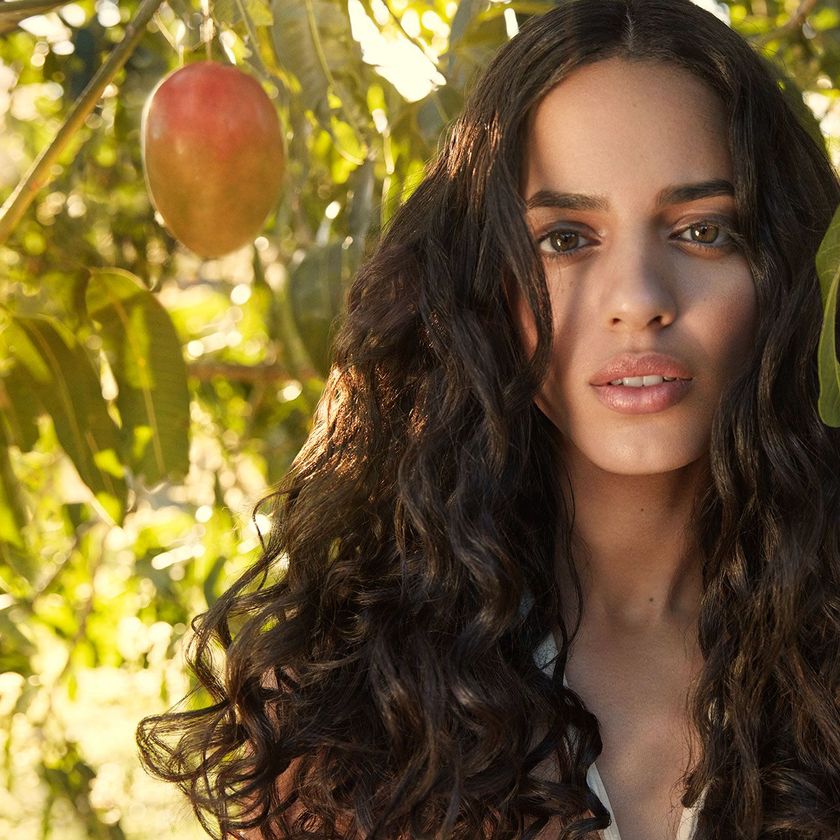 Woman with long dark curly hair standing beside a mango tree in warm sunlight