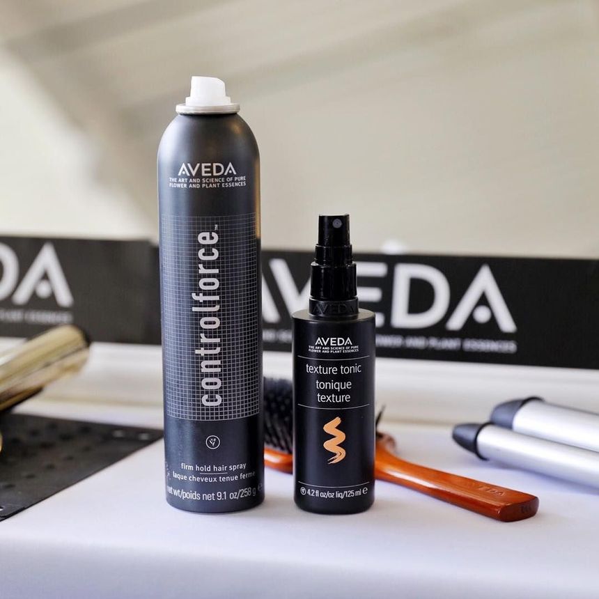 Aveda Control Force hairspray and styling product bottles on a countertop with a comb and laptop in the background