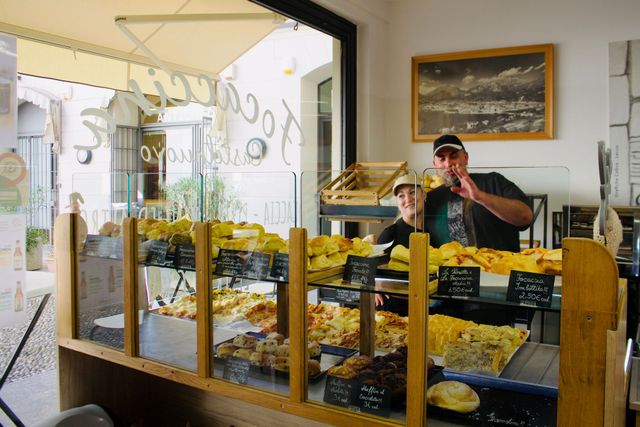 Vetrina di panetteria con prodotti da forno; due persone dietro il bancone, una delle quali fa segno di sì.