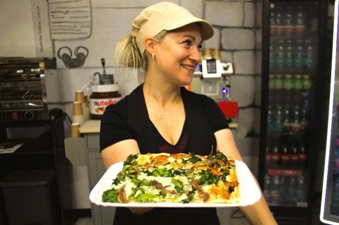 Donna sorridente che tiene in mano un vassoio con una fetta di pizza, nella cucina di un ristorante.