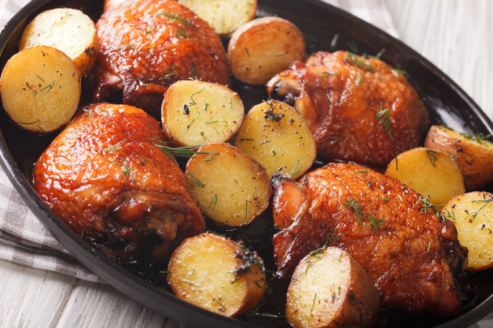 Cosce di pollo arrosto con patate a fette in una teglia nera, condite con erbe aromatiche e salsa