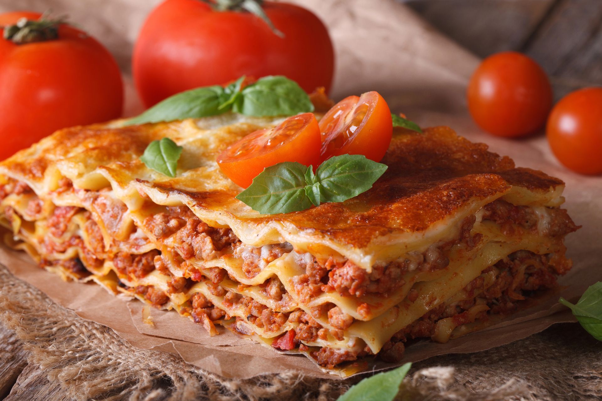 Primo piano di una fetta di lasagna guarnita con basilico, su un tagliere di legno con pomodori sullo sfondo