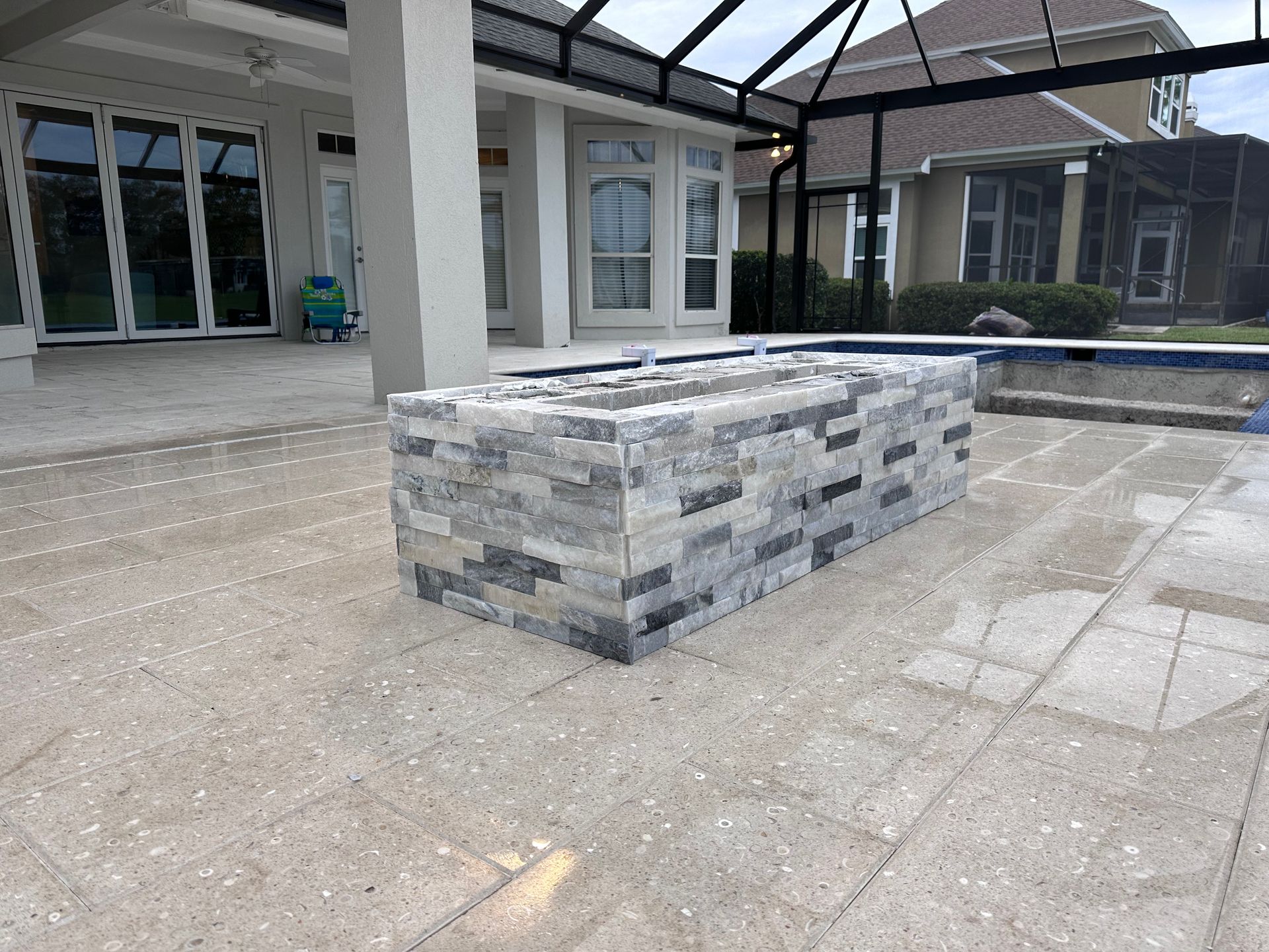 Rectangular stone fire pit on a light-colored patio, with a pool and house in the background.