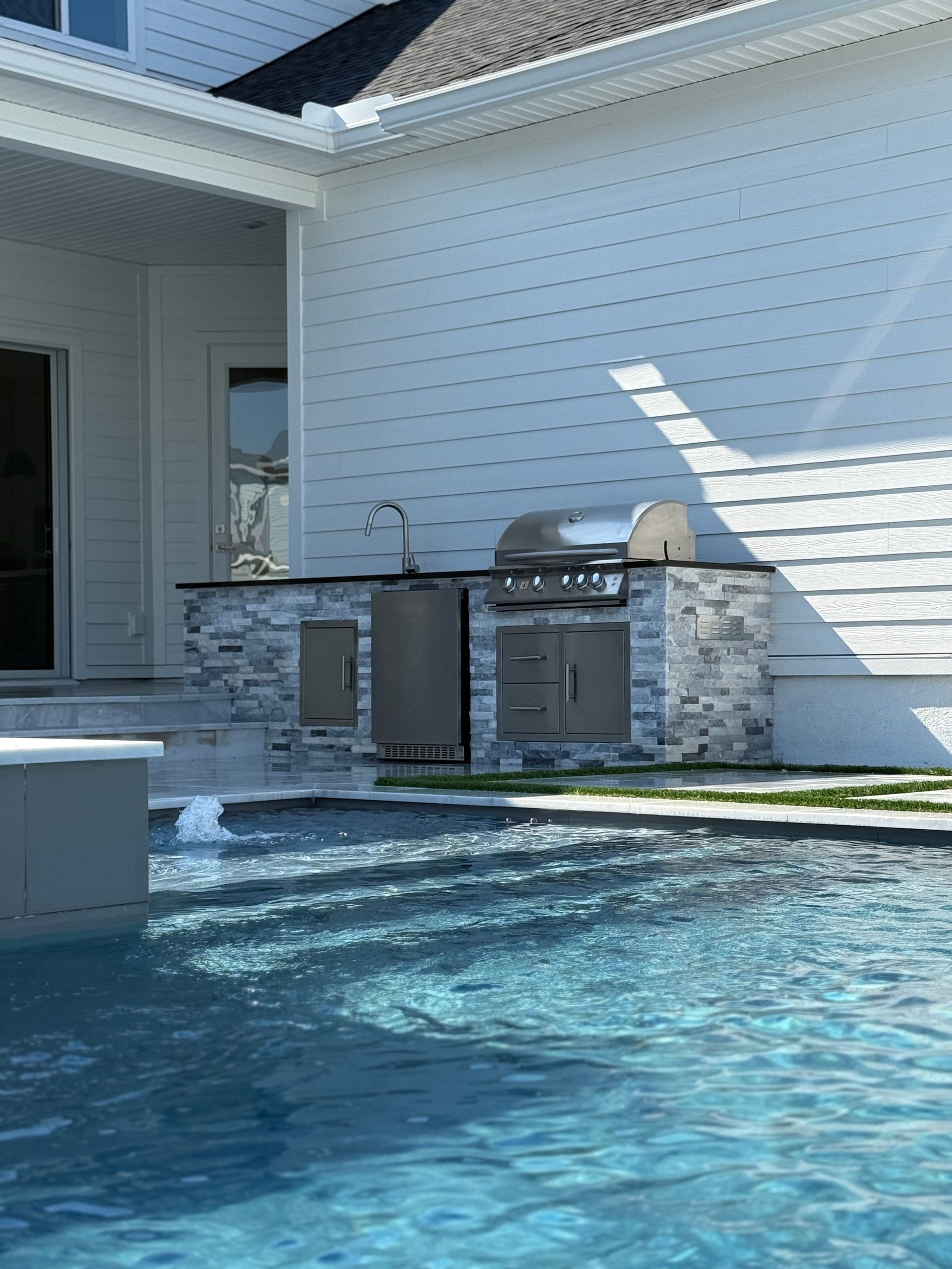 Outdoor kitchen with grill, fridge, and stone facade next to a swimming pool.