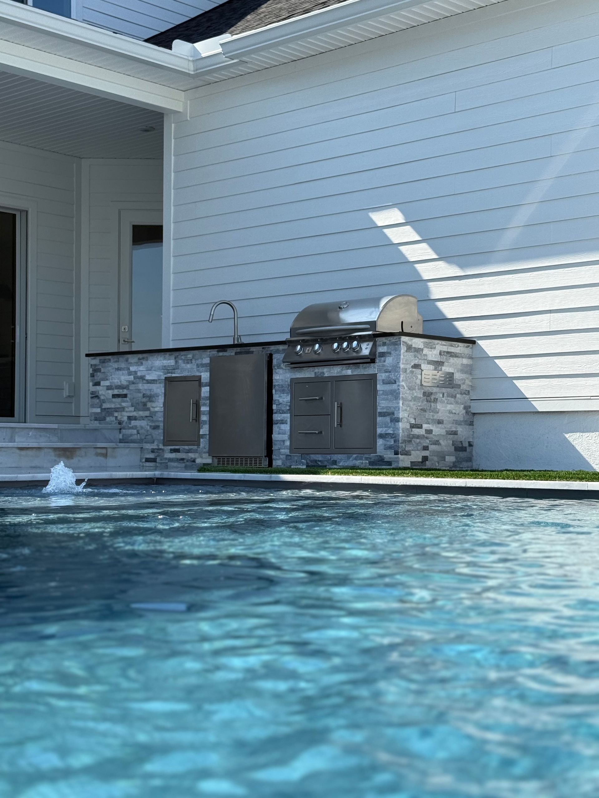 Outdoor kitchen with a grill, sink, and refrigerator next to a swimming pool.