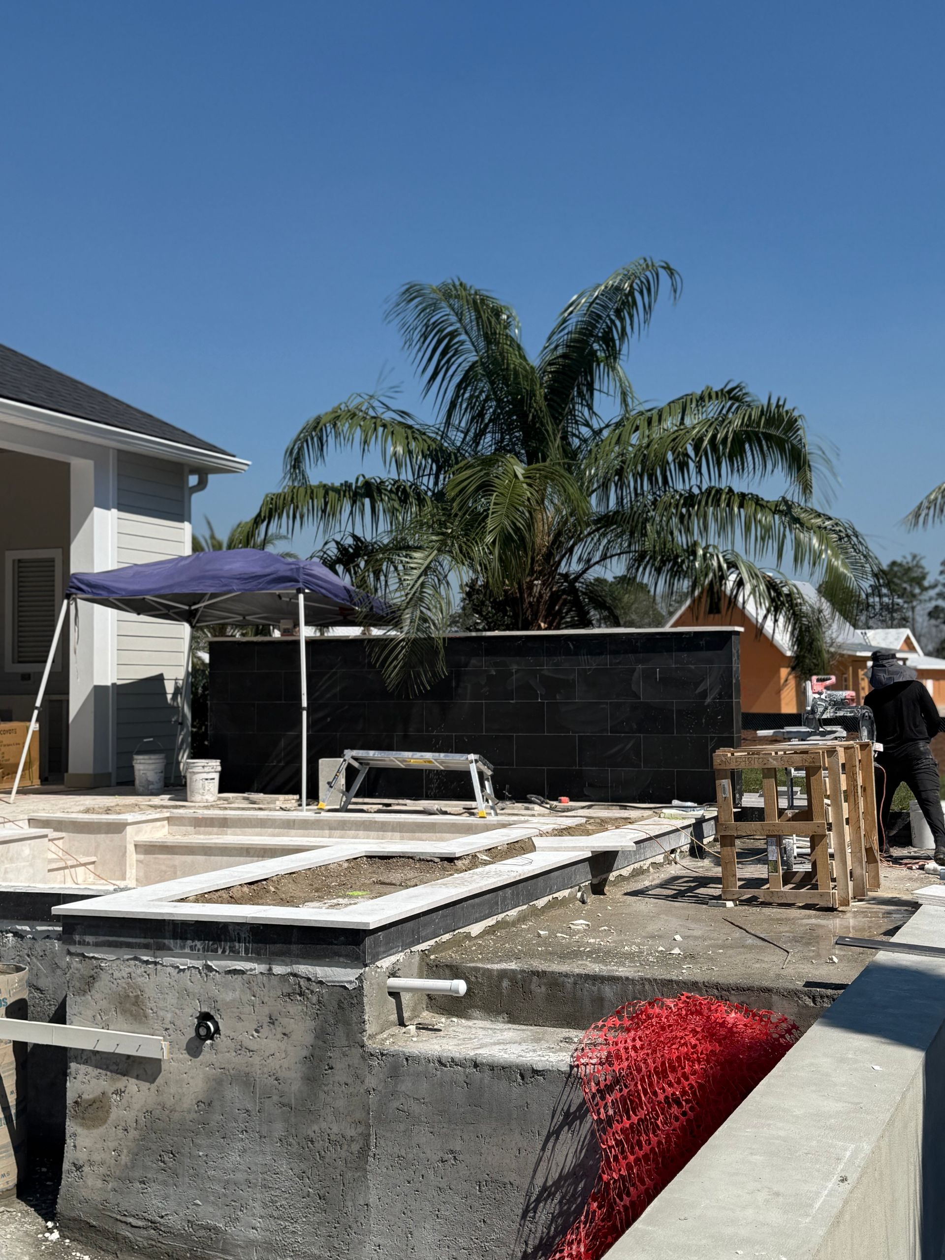 Pool under construction; grey concrete, white tile trim, black wall, palm tree, blue umbrella, sunny day.