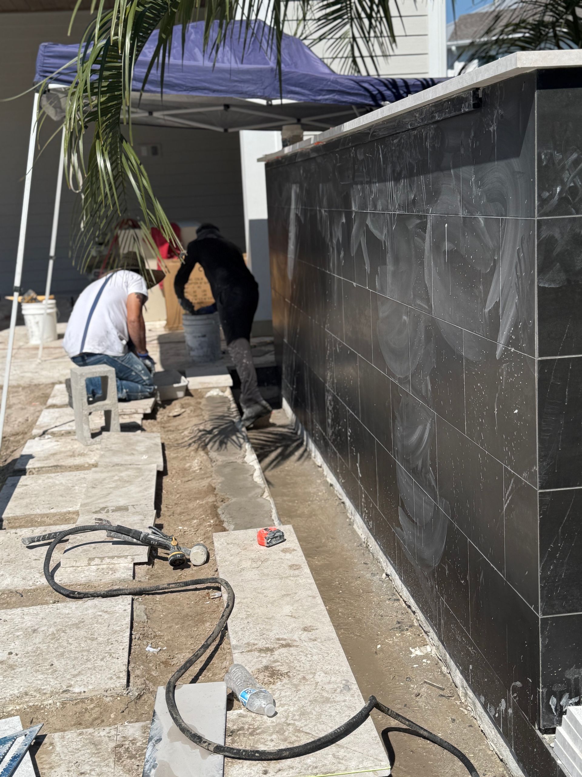 Two workers tiling a black wall outdoors. Construction tools and materials are visible.