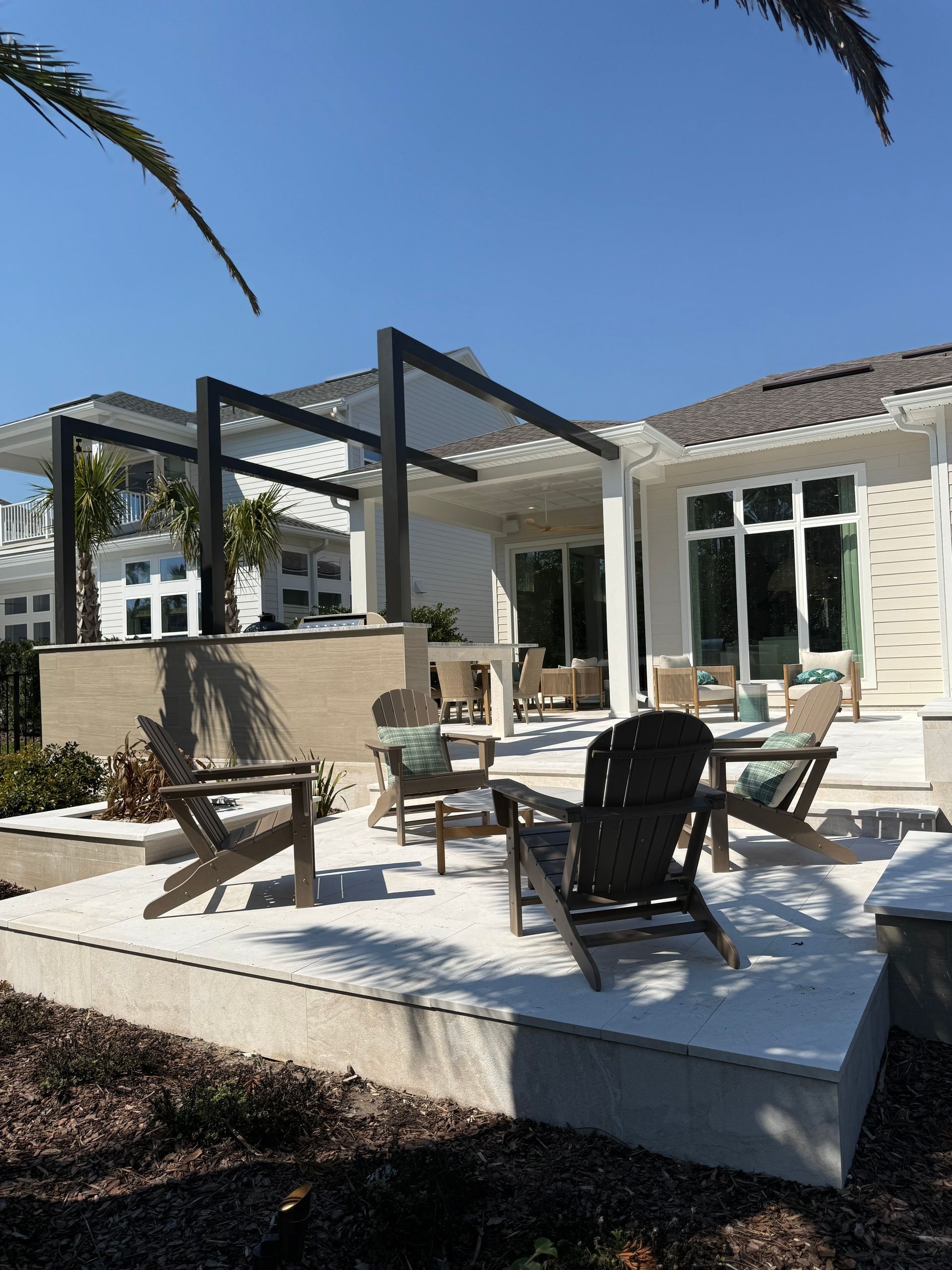 Patio with seating under a black pergola, near a white house under a blue sky.