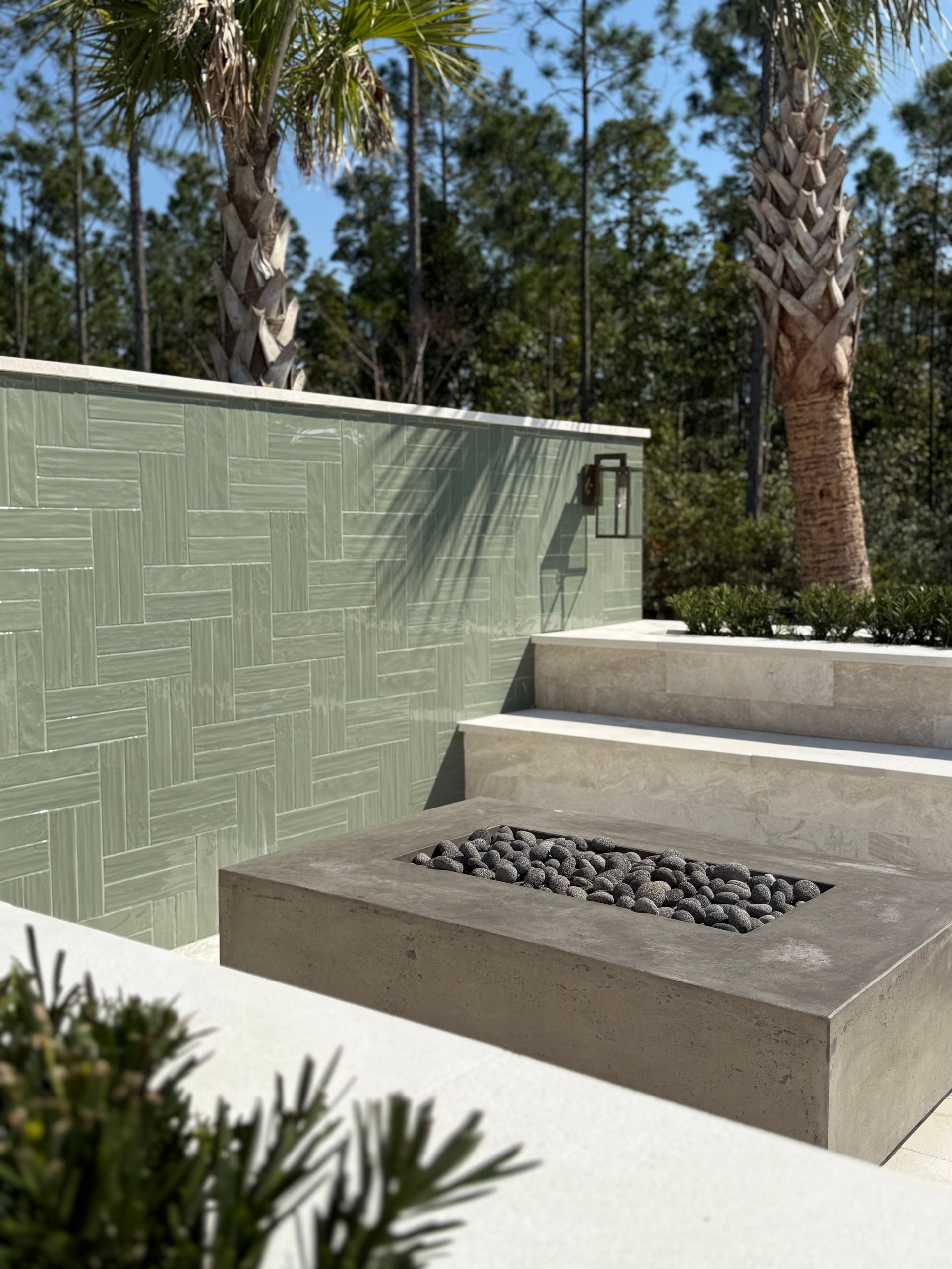 Outdoor concrete fire pit, green tiled wall, steps, palm trees, and greenery.