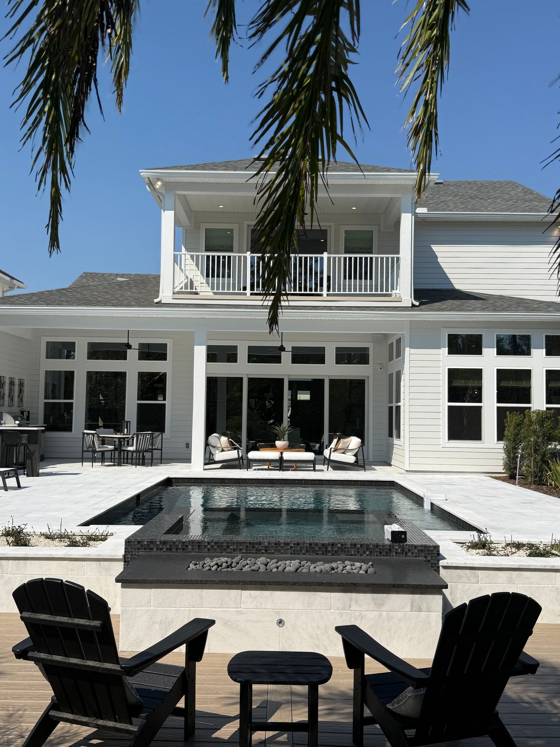 White two-story house with a pool and chairs on a sunny day. Palm fronds frame the view.