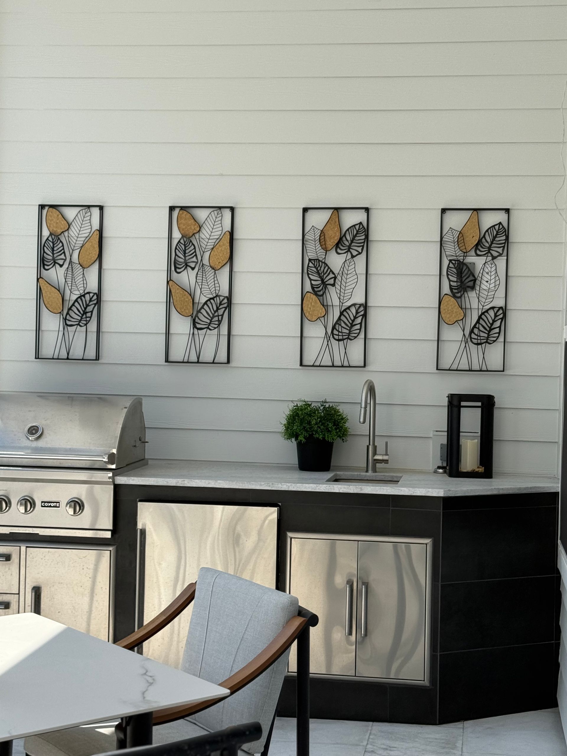 Outdoor kitchen with grill, sink, and artwork on white shiplap wall. Black cabinetry, stainless steel appliances.