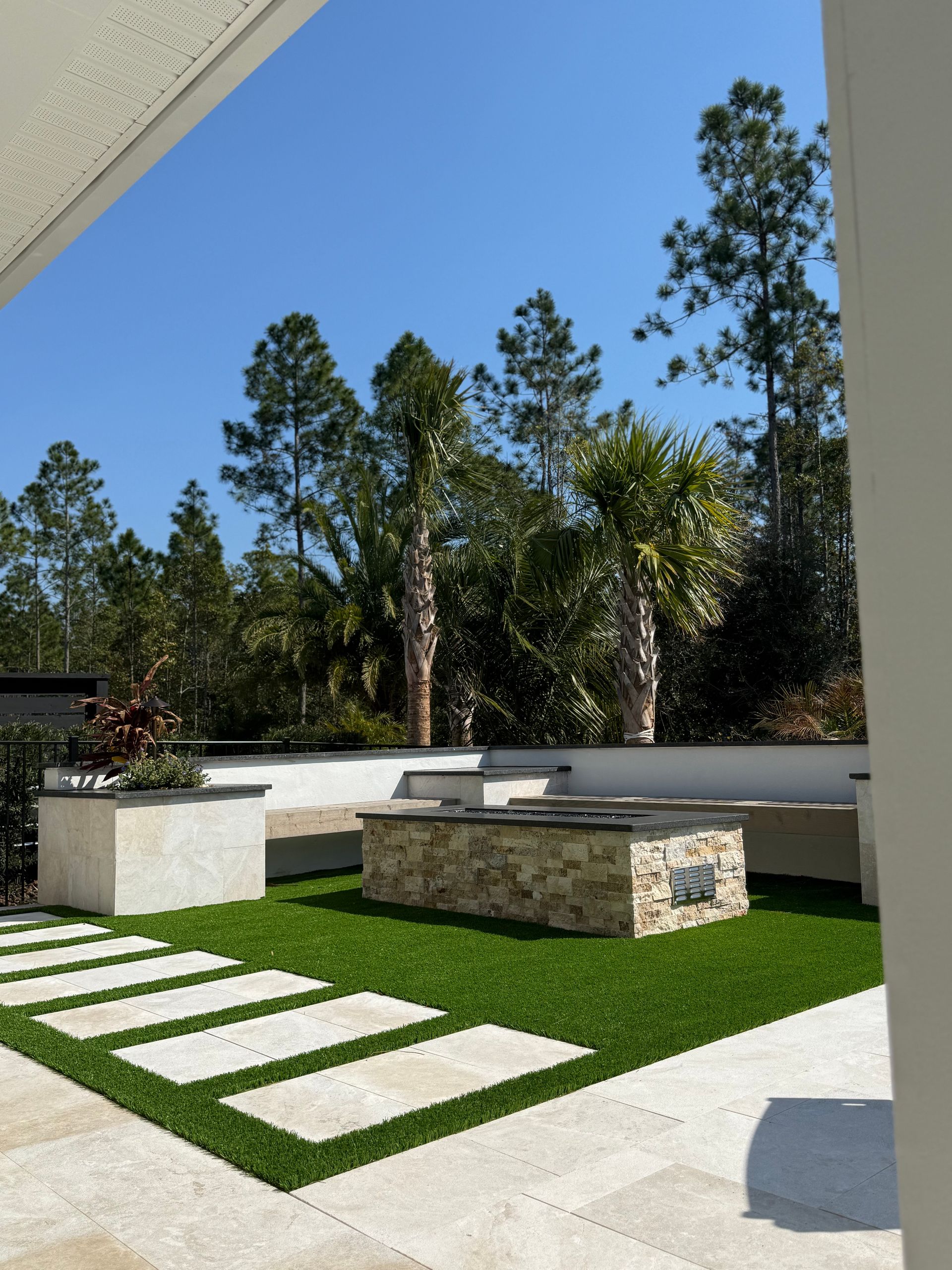 Outdoor patio with stone fire pit, pavers, and lush green artificial turf, set against a backdrop of tall trees and blue sky.