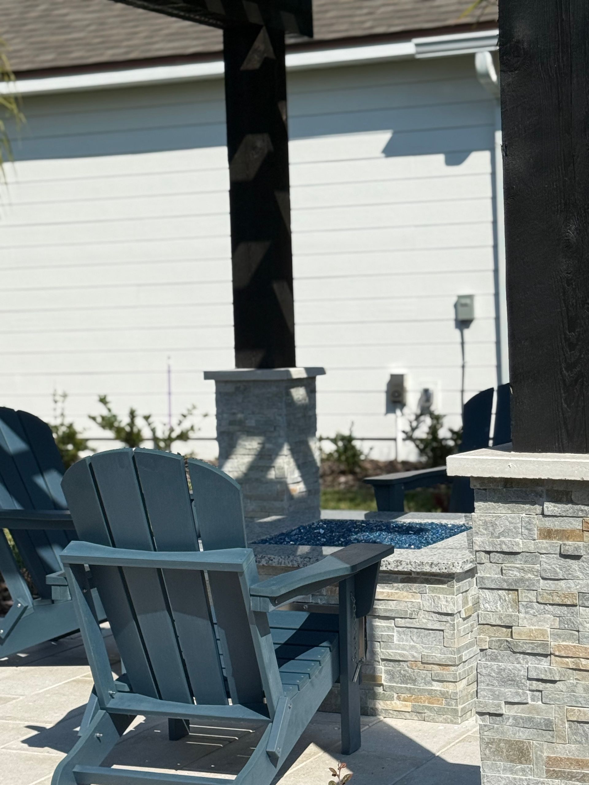 Blue Adirondack chairs around a fire pit on a patio with stacked stone columns and a pergola.