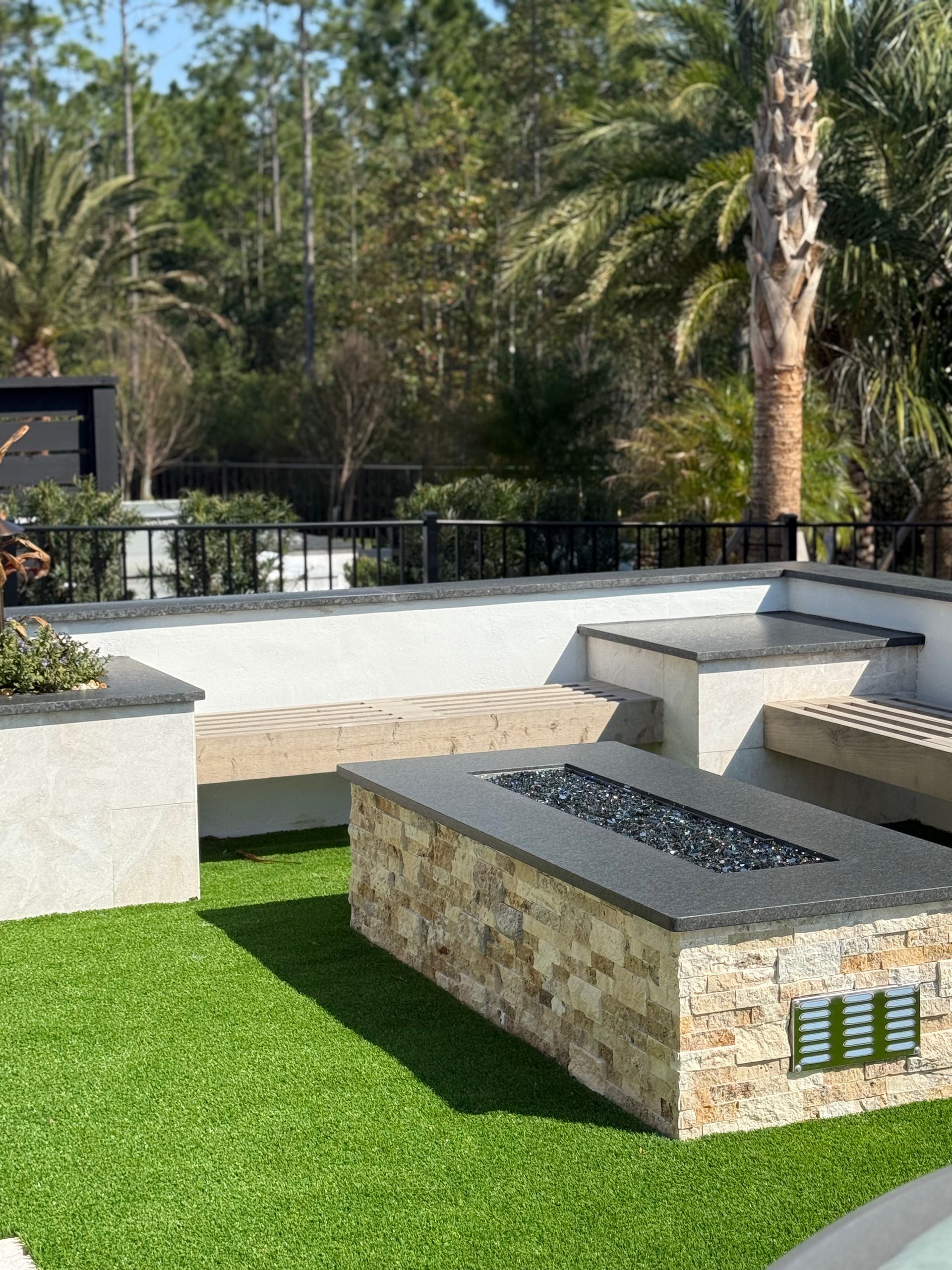 Outdoor patio with a stone fire pit, built-in seating, and green artificial turf. Palm trees and fence in the background.